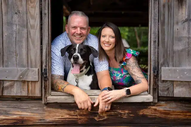 Man and woman smiling with a black and white dog framed in a rustic barn window opening.