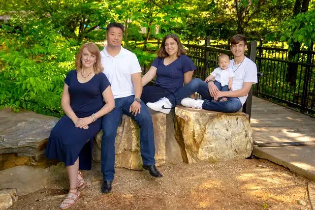 Five-person family group seated on a stone bench in a shaded park setting with green foliage behind them.