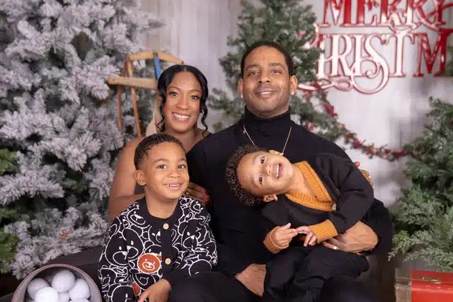 Family of four posing together in front of decorated Christmas trees and holiday signage in studio setting.
