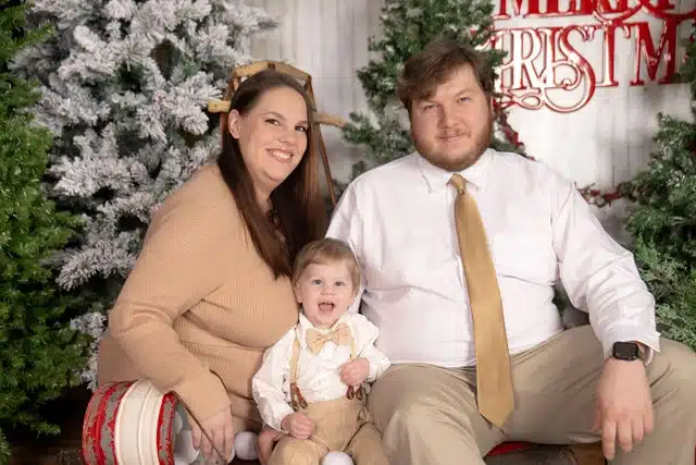 Parents and toddler posed together in front of decorated Christmas trees and garland in studio setting.