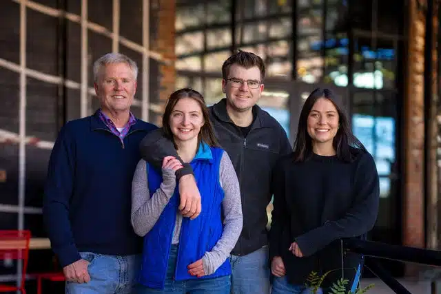 Four people standing together in front of an industrial brick wall with large windows at dusk.