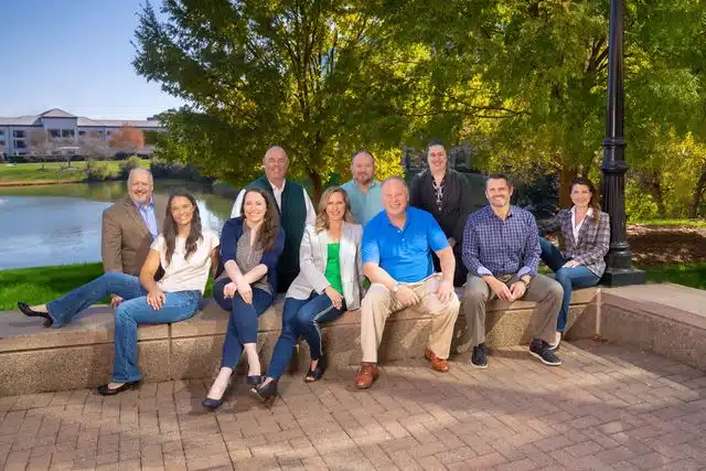 Ten adults seated on a concrete bench beside a pond with trees and buildings in the background.