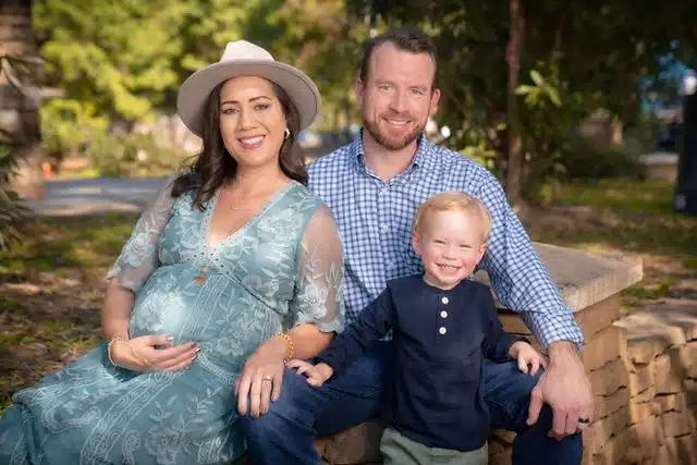 Pregnant woman in blue dress and hat with man and young child seated on outdoor bench surrounded by trees.