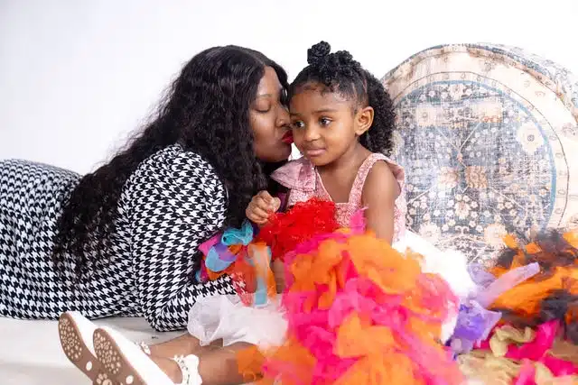 Mother kissing young daughter's cheek while child wears colorful tutu and holds blue toy.