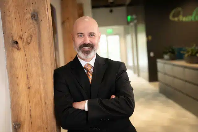 Man in black blazer and striped tie standing with arms crossed against wooden column in modern office.