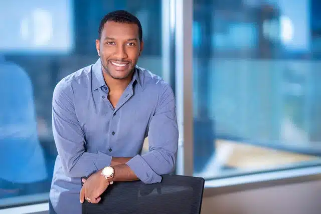 Professional man in lavender button-up shirt leaning against chair with office windows in background.