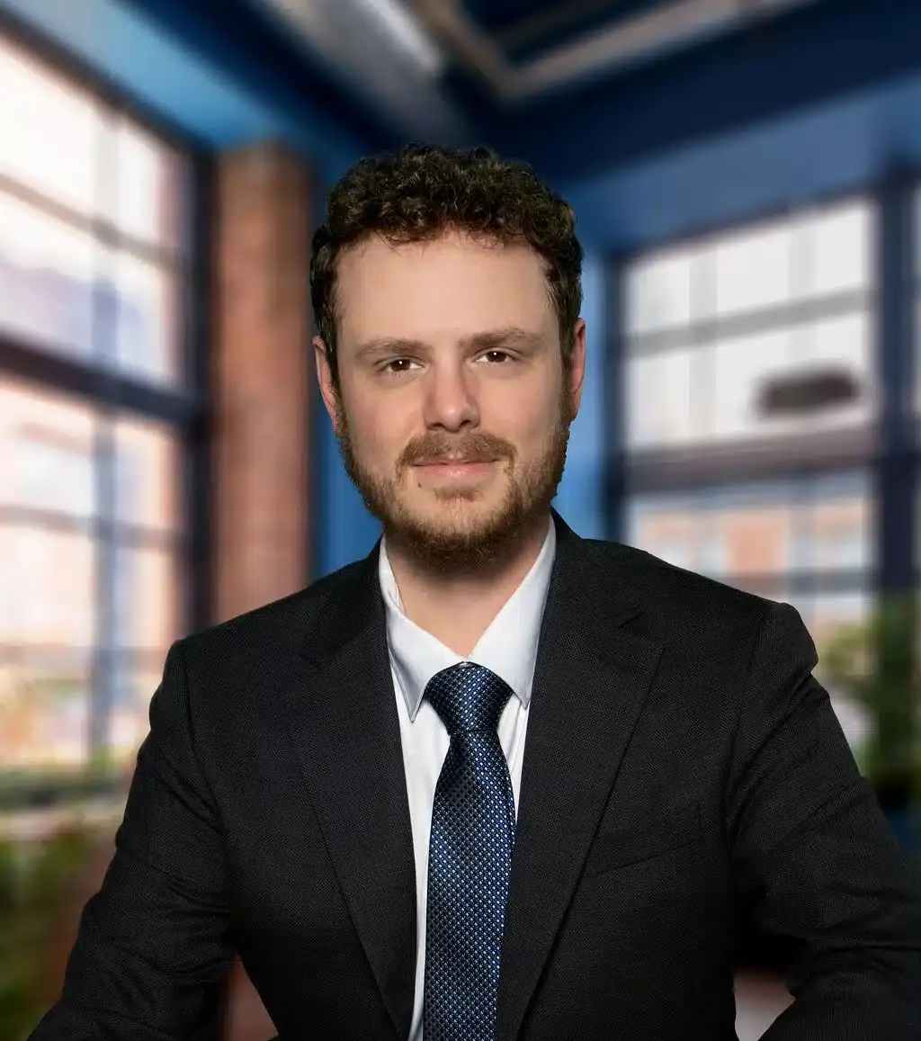 Man in black suit and navy tie seated indoors with blue-toned interior and windows behind him.