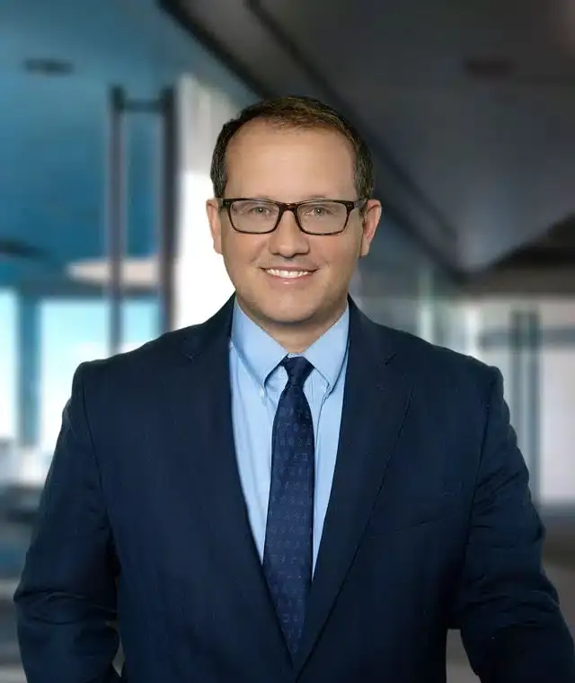 Man in navy blazer and blue tie wearing glasses, smiling at camera in modern office with glass windows.