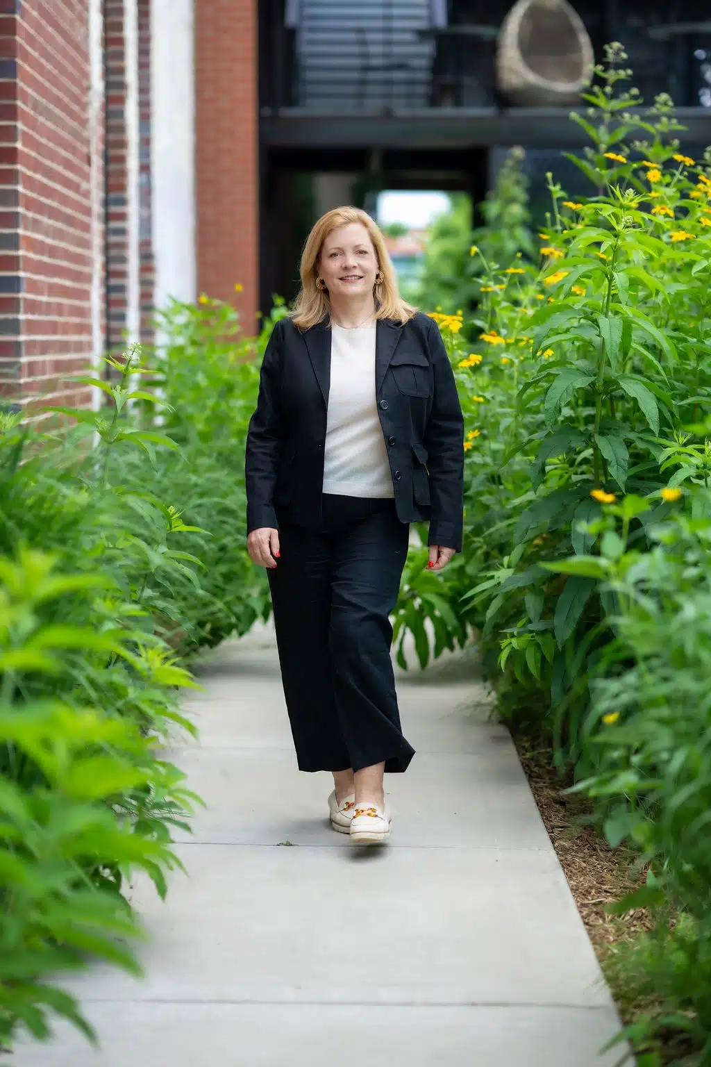 Woman in black blazer and white shirt walking through lush green garden corridor with yellow flowers.
