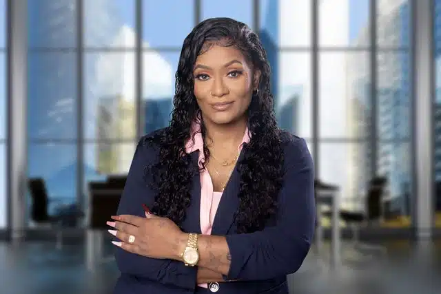 Professional woman in dark blazer and pink blouse posed in front of floor-to-ceiling windows in a high-rise office.
