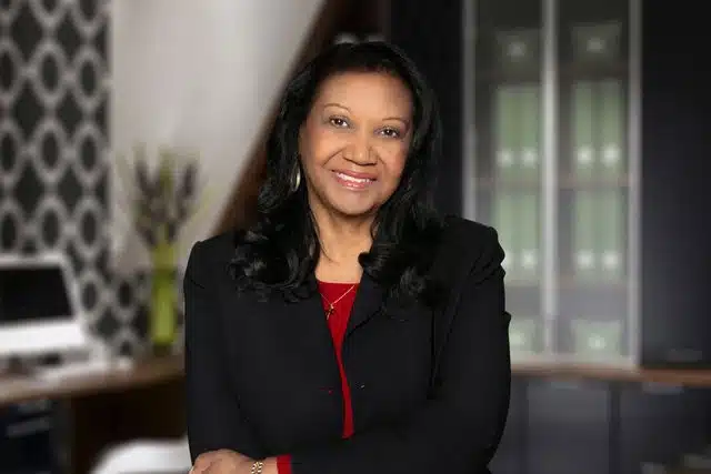 Woman in black blazer and red top smiling at camera in modern office setting with geometric wallpaper.