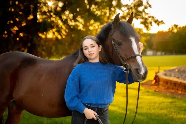Teen girl in blue sweater standing with brown horse in pasture at sunset.