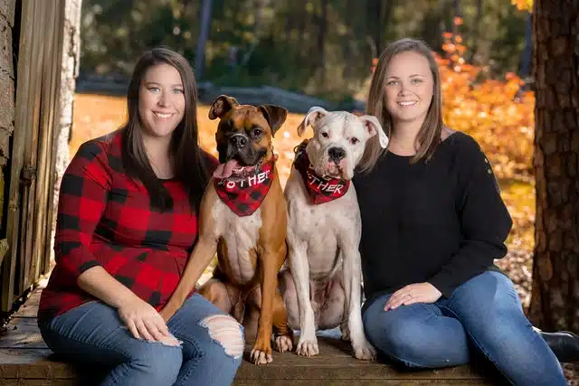 Two women posing with a brown dog and a white dog wearing red bandanas, seated outdoors with autumn foliage in background.