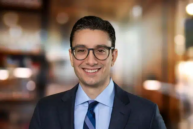 Young man wearing glasses, navy suit jacket, and light blue dress shirt with navy tie, smiling at camera.