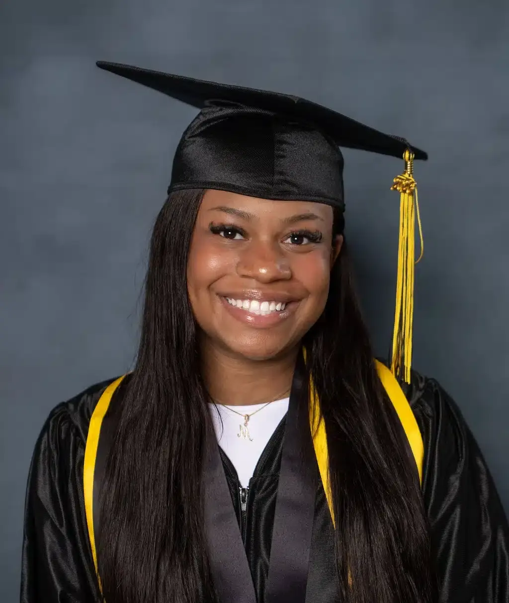 Young woman in black graduation cap and gown with gold tassel, smiling at camera against gray backdrop.