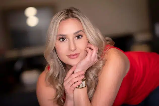 Young woman with blonde wavy hair wearing a red sleeveless top, posed with hand near face.